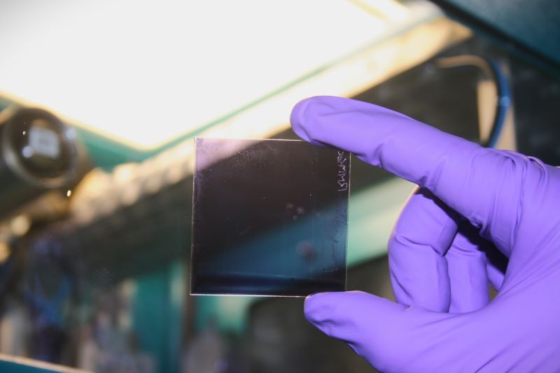 A gloved hand holding a 5cm by 5cm transparent organic solar cell up to the light so you can see through it.